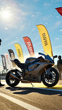 a grey sportbike on a race track with flags and riders under a bright sky