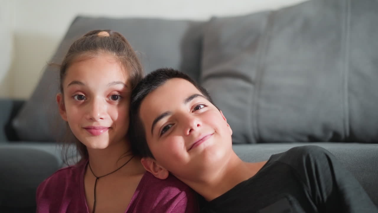 Il fratello minore appoggia la testa sulla spalla della sorella in modo giocoso mentre entrambi guardano la fotocamera con un sorriso gentile, seduti in casa sul pavimento, appoggiati a un divano grigio, mostrando affetto e legame fraterno