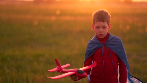 Der-Junge-Im-Kostüm-Eines-Superhelden-Läuft-In-Einem-Roten-Mantel-Und-Lacht-Bei-Sonnenuntergang-Auf-Einer-Sommerwiese-Und-Stellt-Sich-Vor,-Er-Sei-Der-Pilot-Des-Flugzeugs