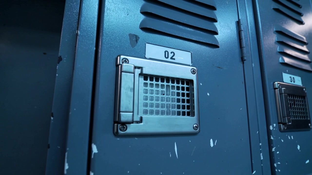 La telecamera si muove a destra, avvicinandosi all'armadietto blu 02 nel corridoio, mostrando la presa d'aria e i graffi.