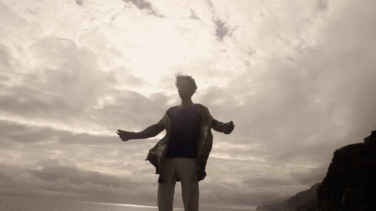 Madeira Island, Portugal - Man Meditating While Standing Under The Clouds With His Clothes Swaying In The Wind In Slow Motion - Low Angle Shot