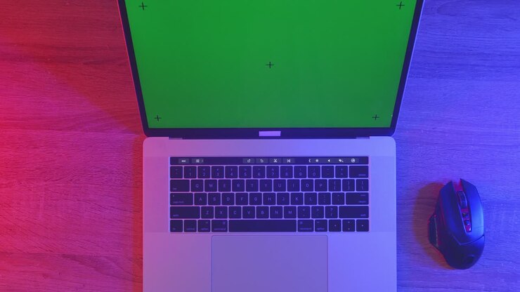 Draufsicht auf einen Laptop-Computer mit Mockup-Greenscreen-Display auf einem Holzschreibtisch, daneben ein Notizbuch mit Stiften, Brille und einem Glas Wasser. Langsames Herauszoomen, Nahaufnahme.