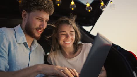A-bearded-guy-in-a-blue-shirt-with-curly-hair-shows-his-girlfriend,-who-is-leaning-on-him-and-laughing,-the-screen-of-his-laptop