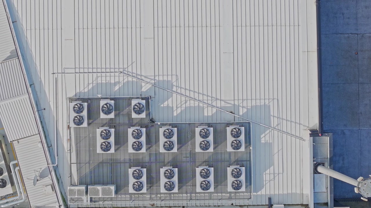 Drone footage captures a bright, top-down view of a data centre rooftop lined with large HVAC units and metallic ventilation. Clean architectural lines, industrial mood, daylight.