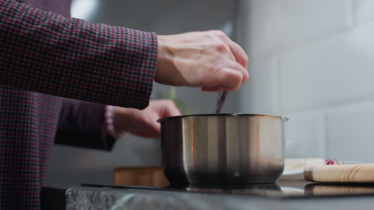 primer plano de sopa agitada a mano en una olla de acero inoxidable en la estufa, con ropa a cuadros, vista parcial de una bandeja de madera con ingredientes en la encimera, entorno de cocina moderno
