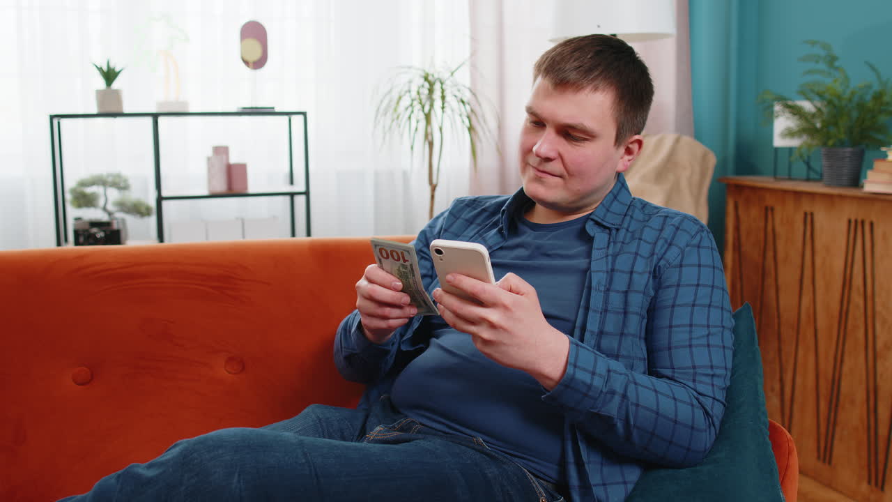 성공한 부자가 스마트폰을 들고 집 소파에 앉아 달러 지폐, 현금을 세고 있다.