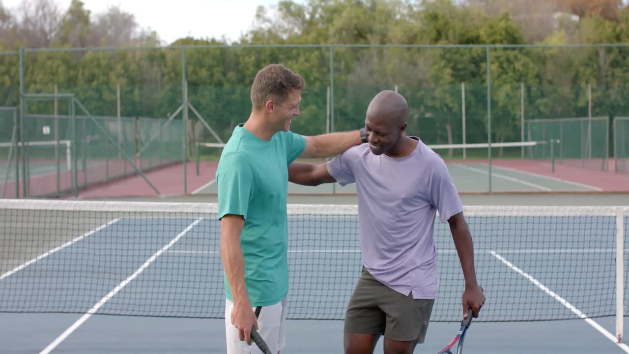 Two happy diverse male friends embracing on outdoor tennis court after game, slow motion