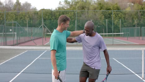 Two-happy-diverse-male-friends-embracing-on-outdoor-tennis-court-after-game,-slow-motion