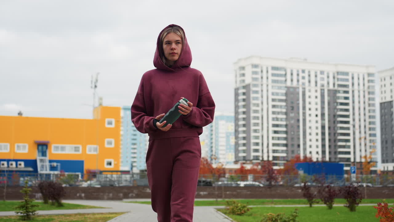 Sport woman in red hoodie holds water bottle while walking on city sidewalk among modern high rise buildings under overcast sky with autumn foliage in casual activewear for urban exercise session