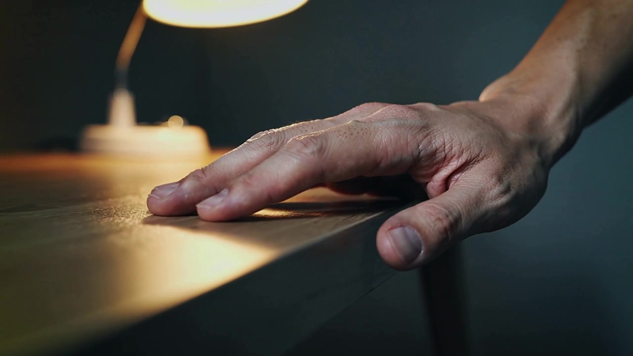 Lowering right hand with bare forearm settling fingers forward on wooden desk near lamp, copy space