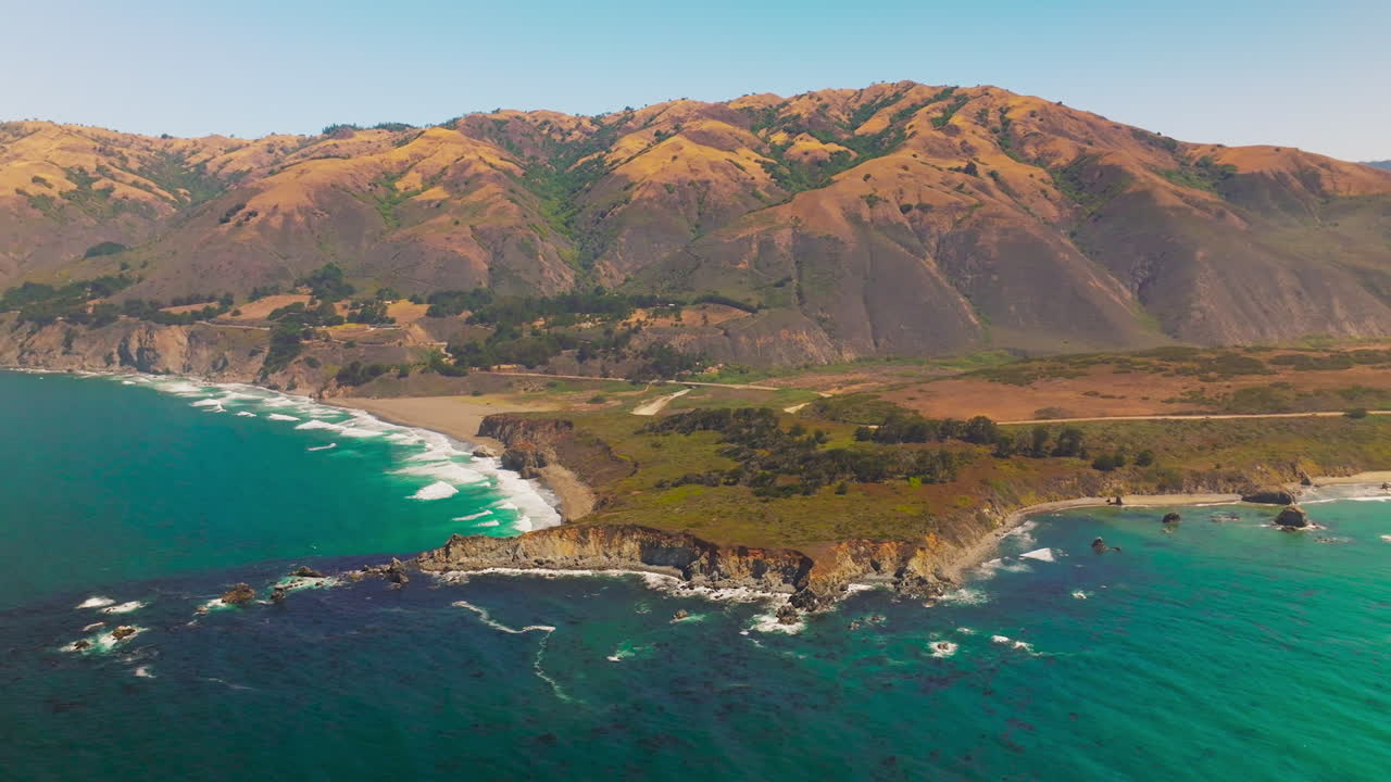 Amazingly beautiful shades of blue in the ocean near rocky shore. Brown orange mountains with rounded tops at backdrop.