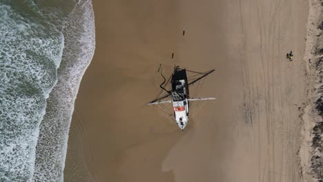 High-drone-view-looking-down-at-a-fishing-trawler-recently-washed-up-on-a-deserted-beach-with-waves-breaking