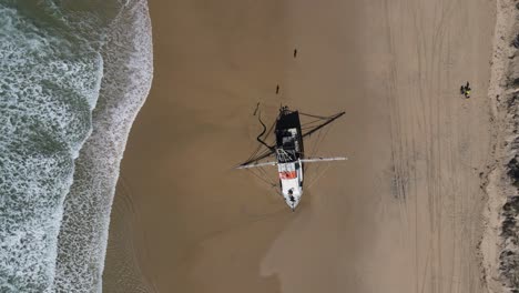 alta vista de drones mirando hacia abajo a un arrastrero de pesca recientemente varado en una playa desierta con olas rompiendo