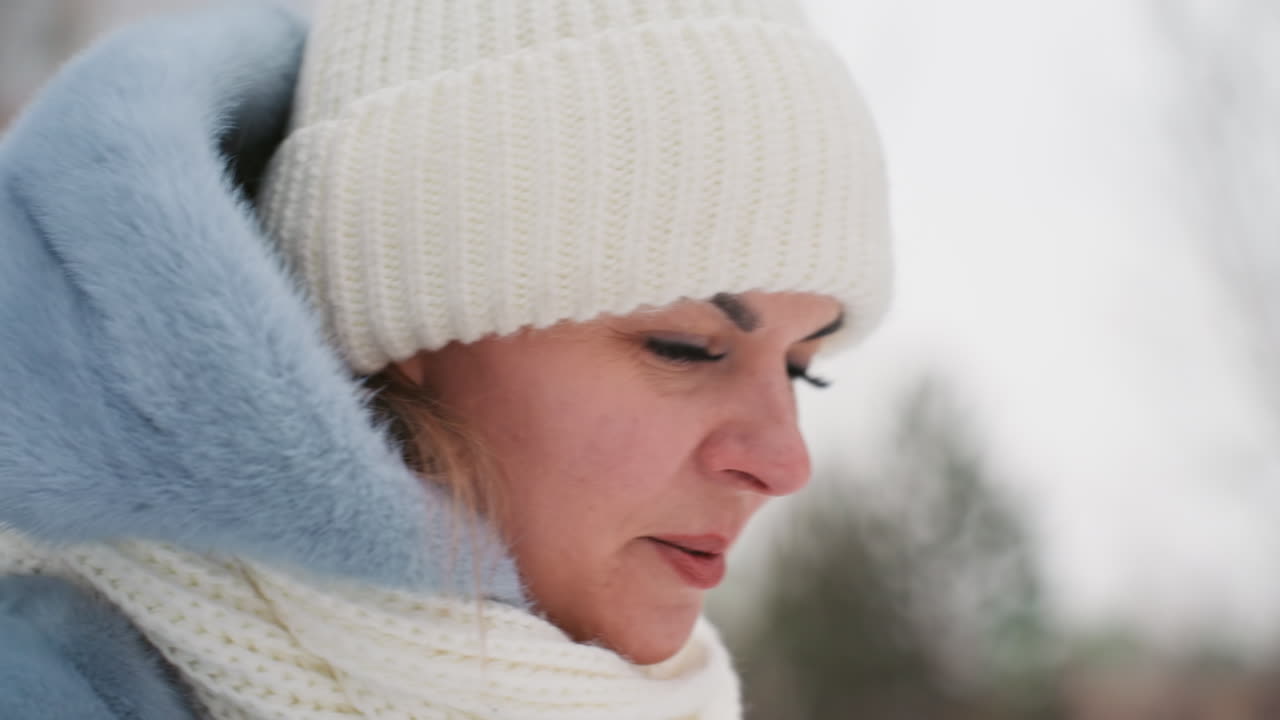 Mamma con berretto di lana bianco in una passeggiata invernale, primo piano pensieroso di profilo, cappotto azzurro e sciarpa color crema, respiro visibile nell'aria fredda, alberi innevati sfocati e cielo nuvoloso e tenue, umore calmo e riflessivo.