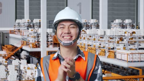 close up of asian male engineer with safety helmet standing in the warehouse with shelves full of delivery goods. smiling to the camera and clapping hands in the storage