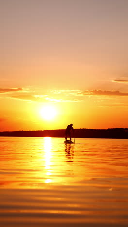 man rows with an oar standing on the sup board. beautiful river reflecting orange light of sunset with lonely sportsman practicing paddle boarding. low angle view. vertical video.