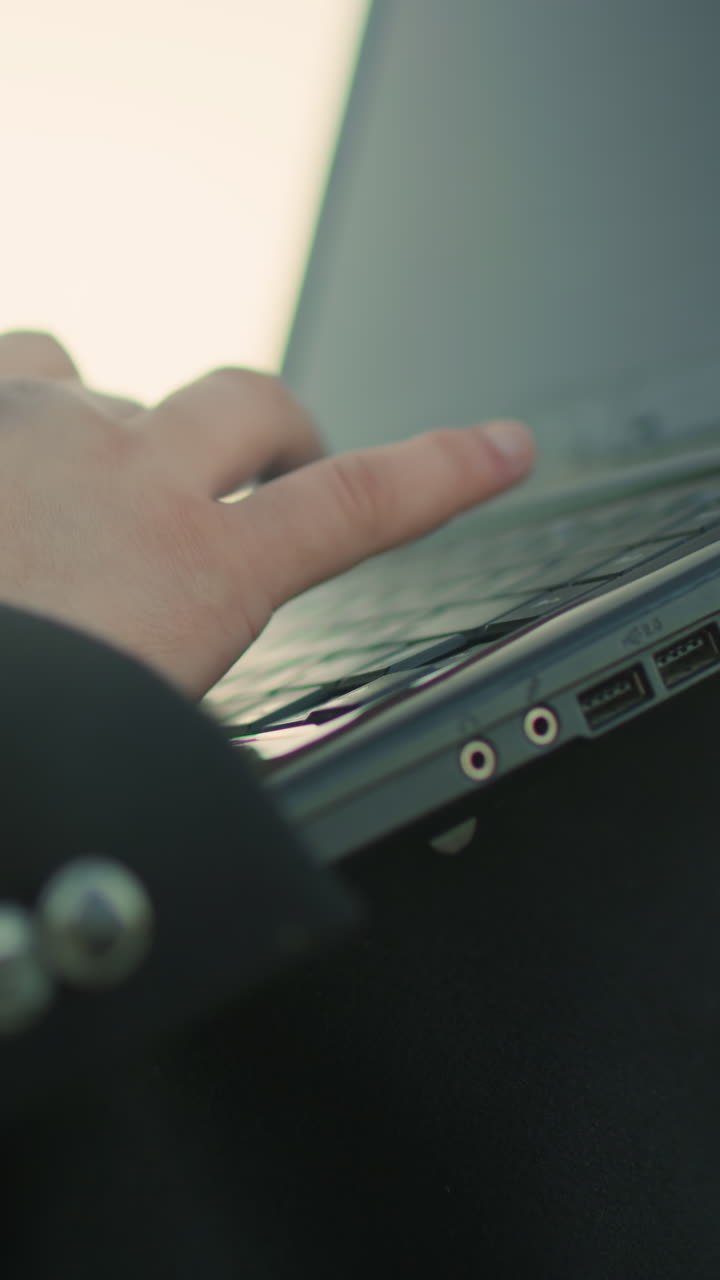 흑색 의복을 입은 개인의 손이 야외에서 노트북을 작동하고 초점과 정확성을 강조합니다. 세부 사항에는 의복 버튼, 노트북 포트 및 부드러운 보케 배경이 포함됩니다.