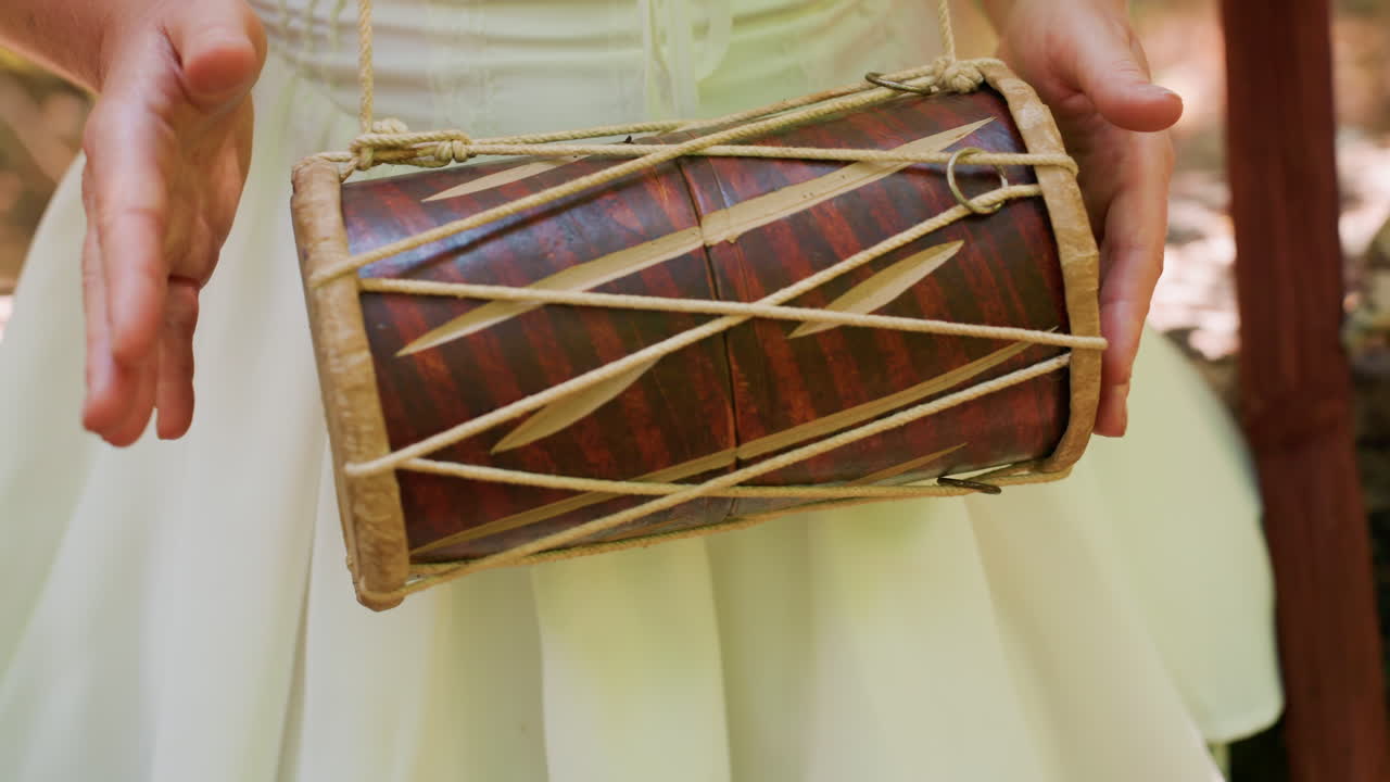 Close up of woman hands rhythmically tapping talking drum, sunlight glimmering through forest canopy, warm tones highlighting wood and rope details
