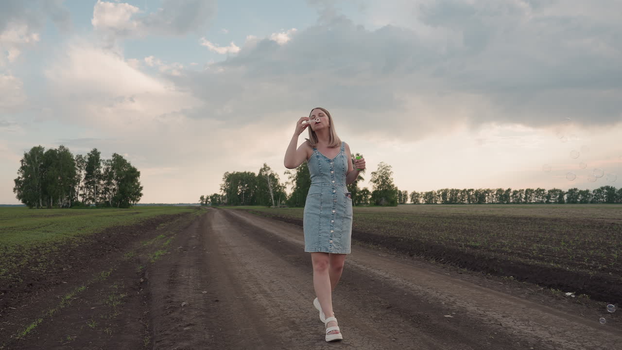 young woman walking along dirt road blowing bubbles through plastic wand under dramatic cloudy sky over vast farm field during golden hour