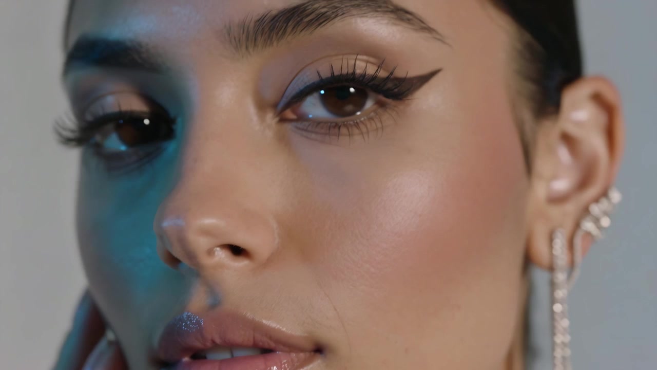 Inquadratura che si allontana, modella che abbassa lo sguardo e si tocca la guancia in studio, mostrando eyeliner alato e orecchino