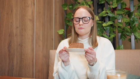 girl with glasses and white sweater holds dessert plate, inhales aroma of chocolate cake with smile while seated at table in warm cafe setting, smoothie and notebook placed on wooden table nearby