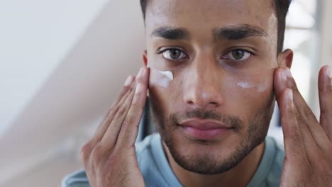 Smiling-biracial-man-looking-in-mirror-and-applying-face-cream-under-eyes-in-bathroom,-slow-motion