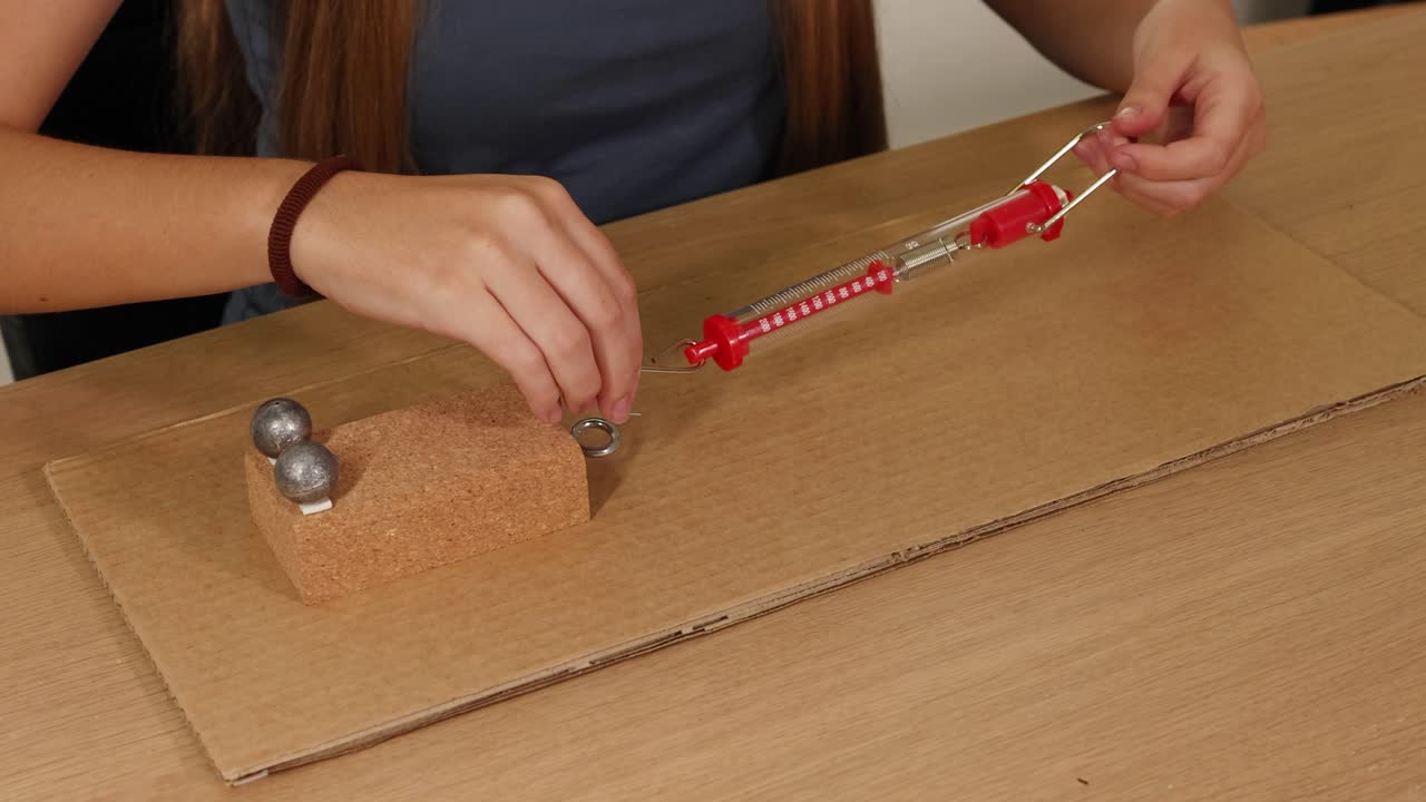 Hands manipulate a spring scale to measure force on a weighted block on cardboard, demonstrating friction principles