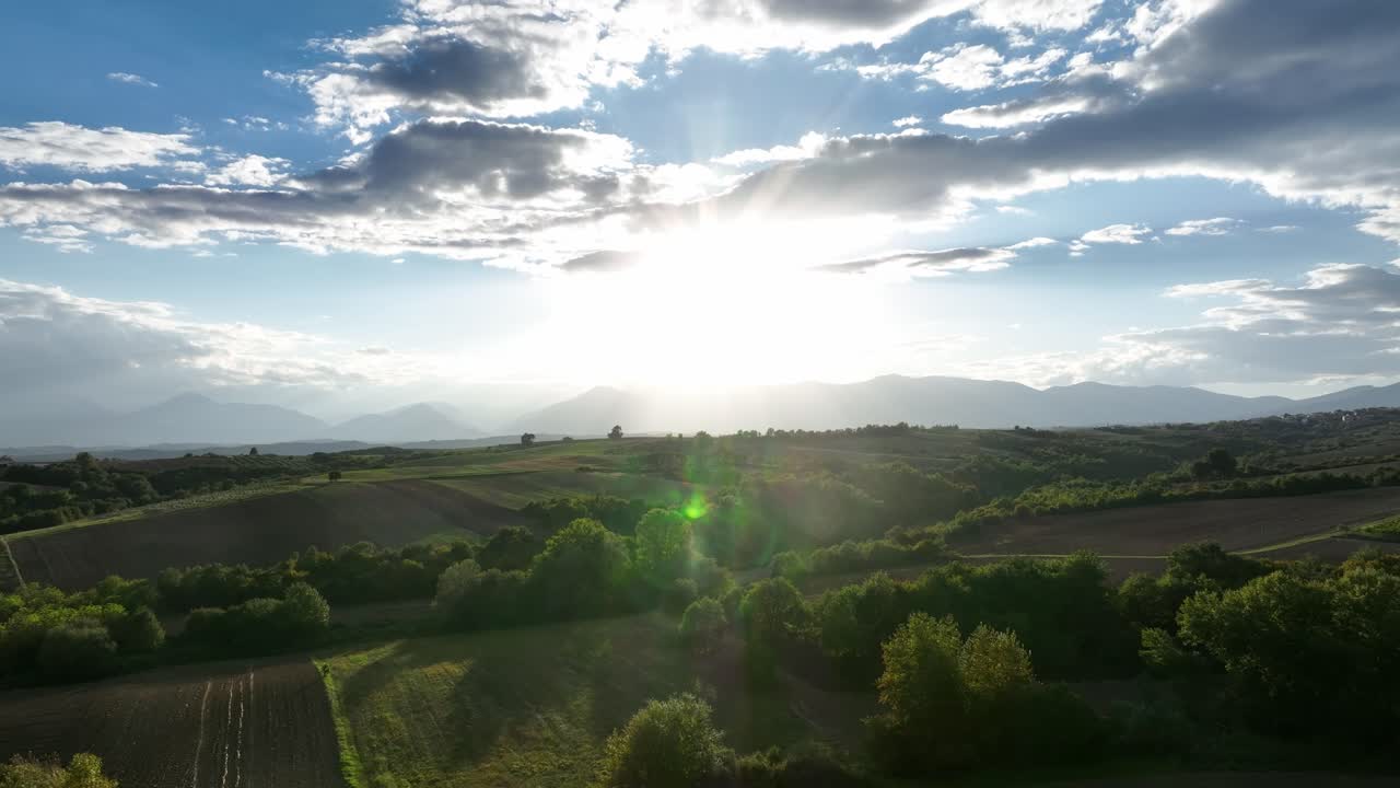 Aerial view of Greece countryside at sunset, sun flare over scenic hills and farmland with clouds in sky, natural beauty, serene vibes and breathtaking landscape.