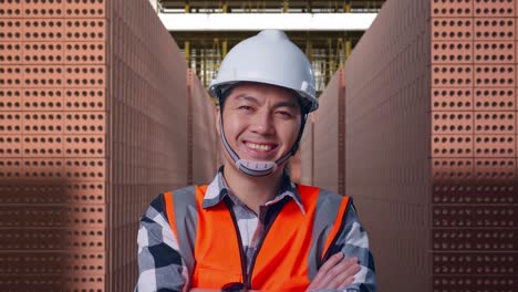 Close Up Of Asian Male Engineer With Safety Helmet Crossing His Arms And Smiling To Camera While Standing With Red Brick Packed in Stacks Are Stored