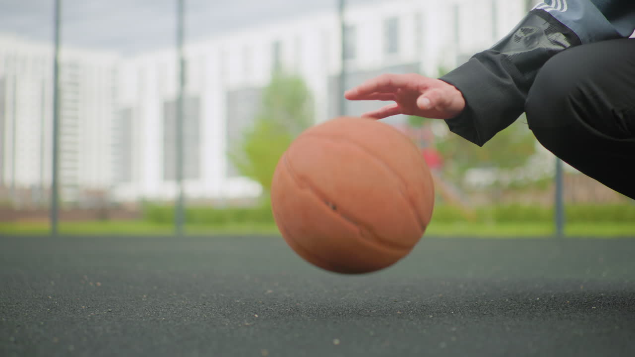 closeup urban basketball dribble on asphalt, hand guiding ball while kneeling in jacket near fenced city court with apartment buildings in soft focus rhythmic bounce, textured rubber surface, subdued