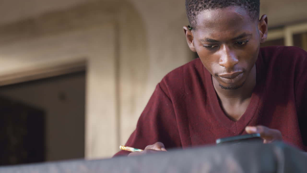 Black artist studying phone reference while sketching with pencil on work surface, leaning over table near doorway, focused expression, closeup portrait, natural light, thoughtful mood, creative