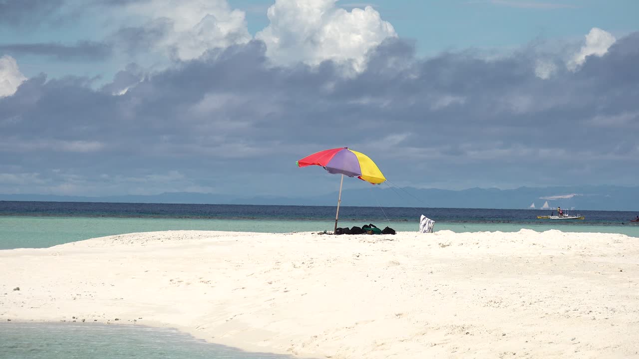 sombrilla de playa roja, azul y amarilla que sopla en el viento en una isla tropical de playa de arena blanca mientras los botes de bomba filipinos pasan bajo un cielo azul nublado