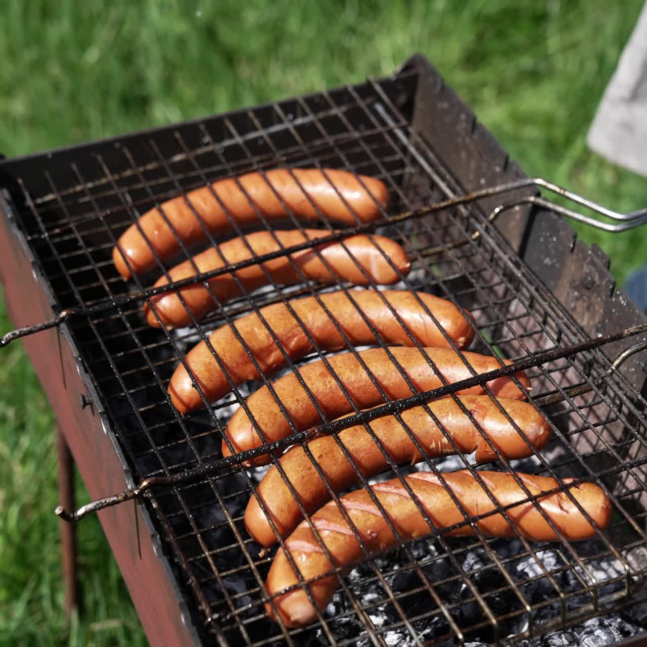 Grigliare salsicce sulla piastra. Grigliare cibo all'aperto. Salsicce calde che arrostiscono nel fumo. Barbecue su sfondo di erba verde