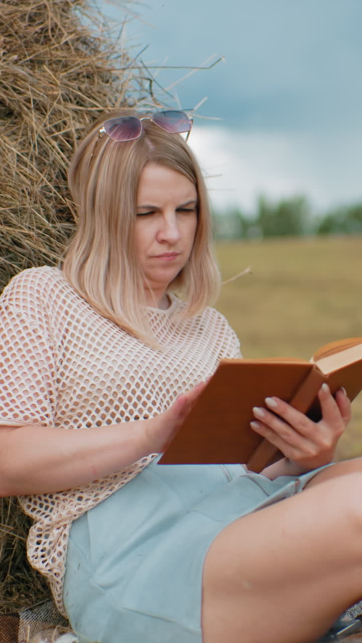joven sentada en una manta, descansando contra una bala de heno, volviendo las páginas del libro reflexivamente, las piernas cruzadas, un cuaderno verde a su lado en la hierba, el fondo borroso del campo con árboles y balas de heno