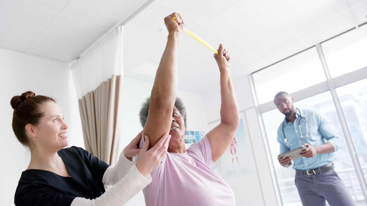 Diverse senior woman exercising with band and female doctor advising in hospital, slow motion