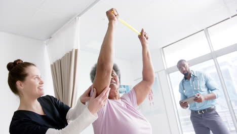 Diverse-senior-woman-exercising-with-band-and-female-doctor-advising-in-hospital,-slow-motion