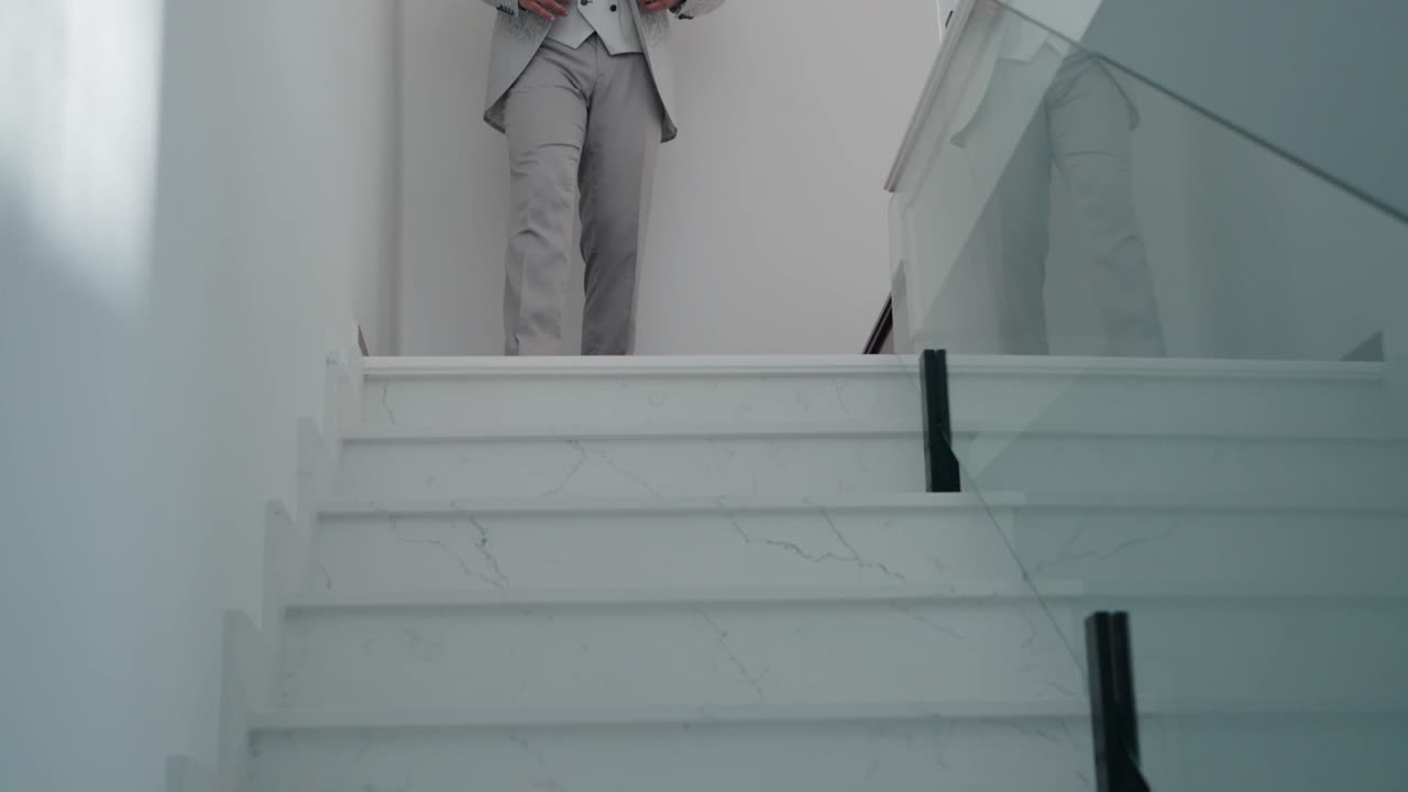 Elegant groom in a light gray suit walking down white marble stairs before the ceremony