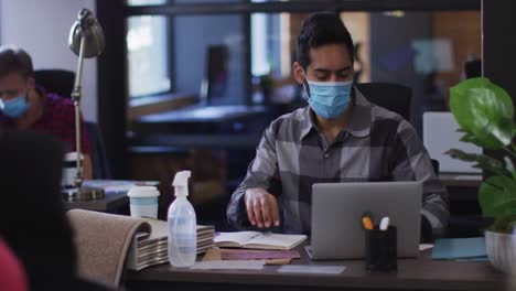 middle eastern man wearing face mask taking notes and using laptop at modern office