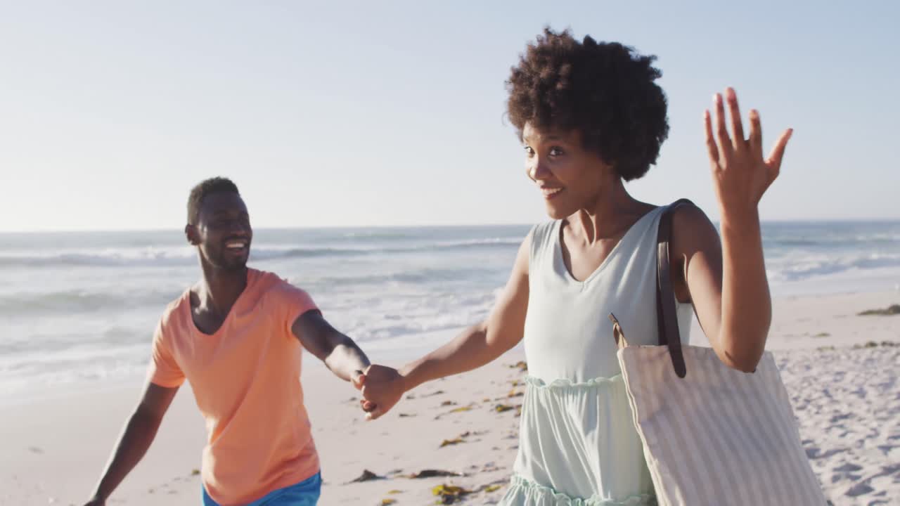 Smiling african american couple holding hands and walking on sunny beach