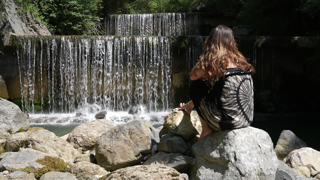 Woman sitting on rocks watching a peaceful waterfall near Walensee lake in Switzerland