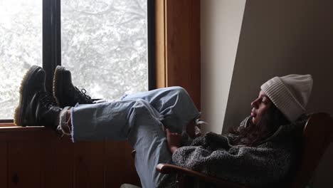 a young woman sits inside with her feet on the windowsill, admiring the snow outsides