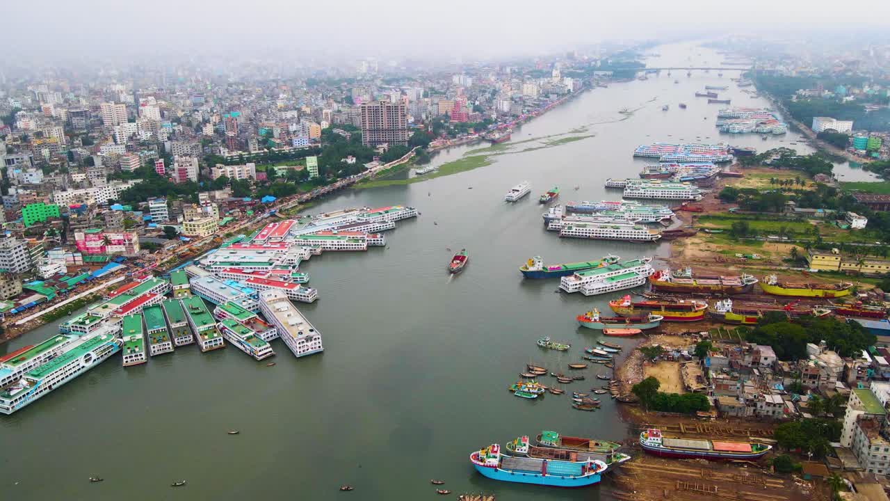 Ripresa aerea del terminal navale della città di Dhaka sul porto fluviale di Buriganga, che mostra numerose navi e lo skyline della città, con un cielo fosco a causa dell'inquinamento.