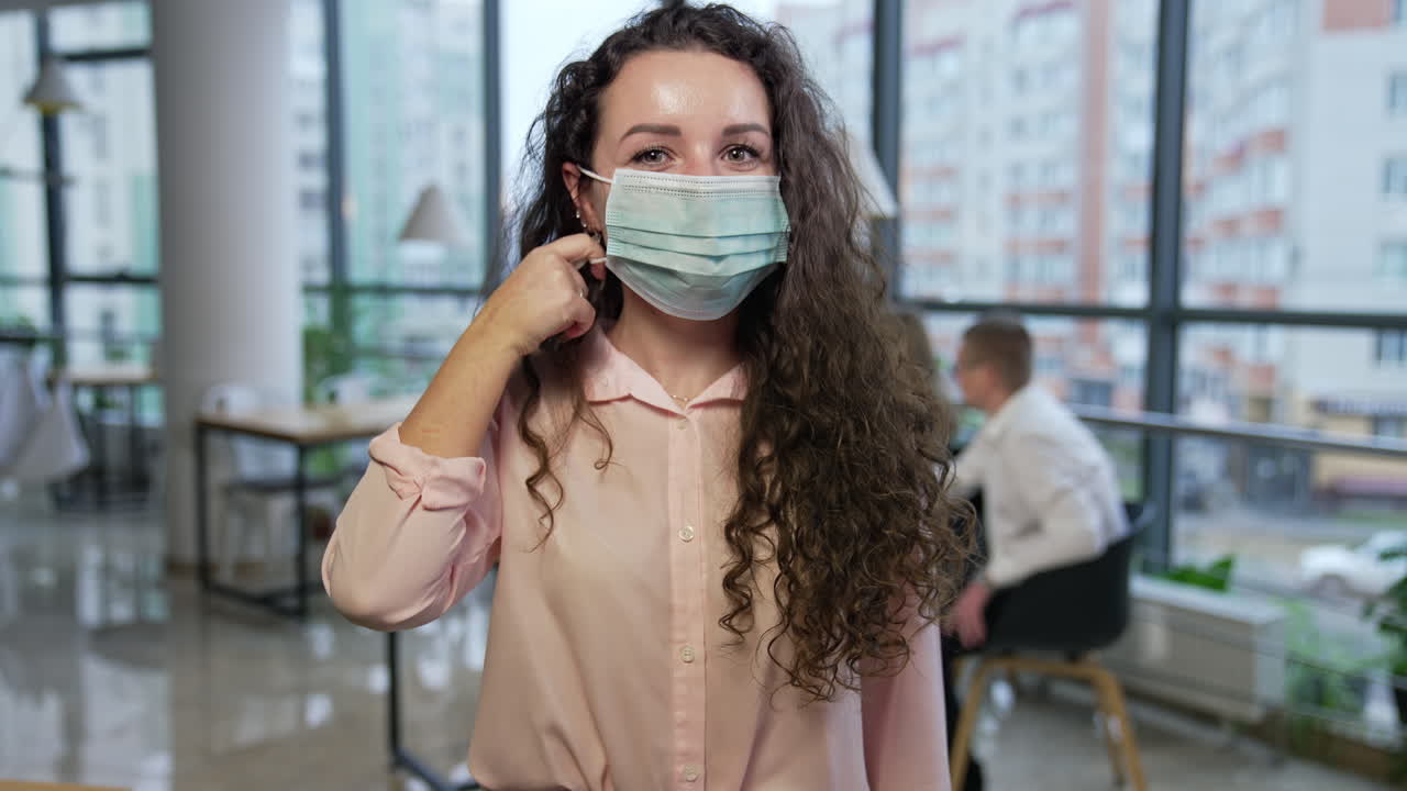 Dark-haired woman wearing mask in office. Girl showing her fingers at the mask, takes it off and smiles cheerfully. Happy lady holding a mask in her hand by the face.