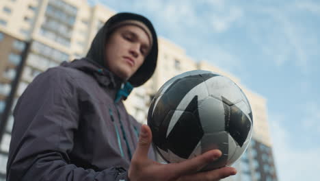 man expertly spinning soccer ball on palm with focused expression, dynamic angle emphasizes skill, blurred urban high-rise background and mesh bar