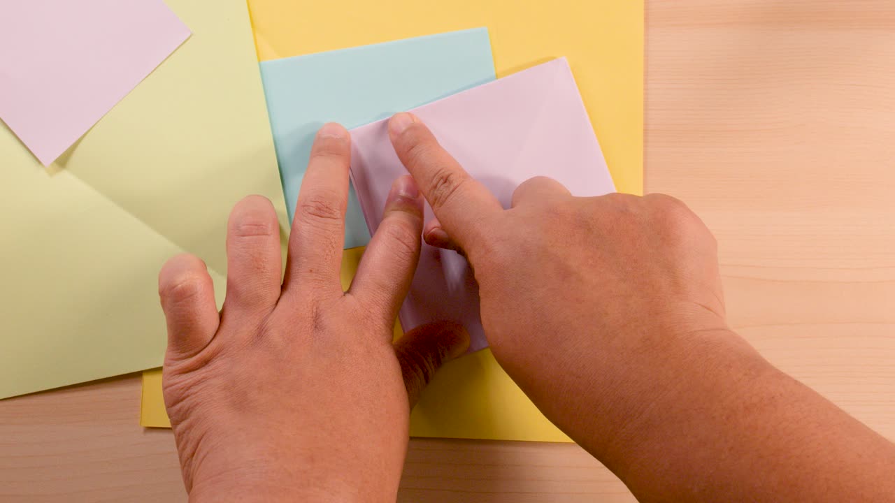 Adult hands carefully fold pastel origami paper, overhead view, soft lighting, steady camera angle