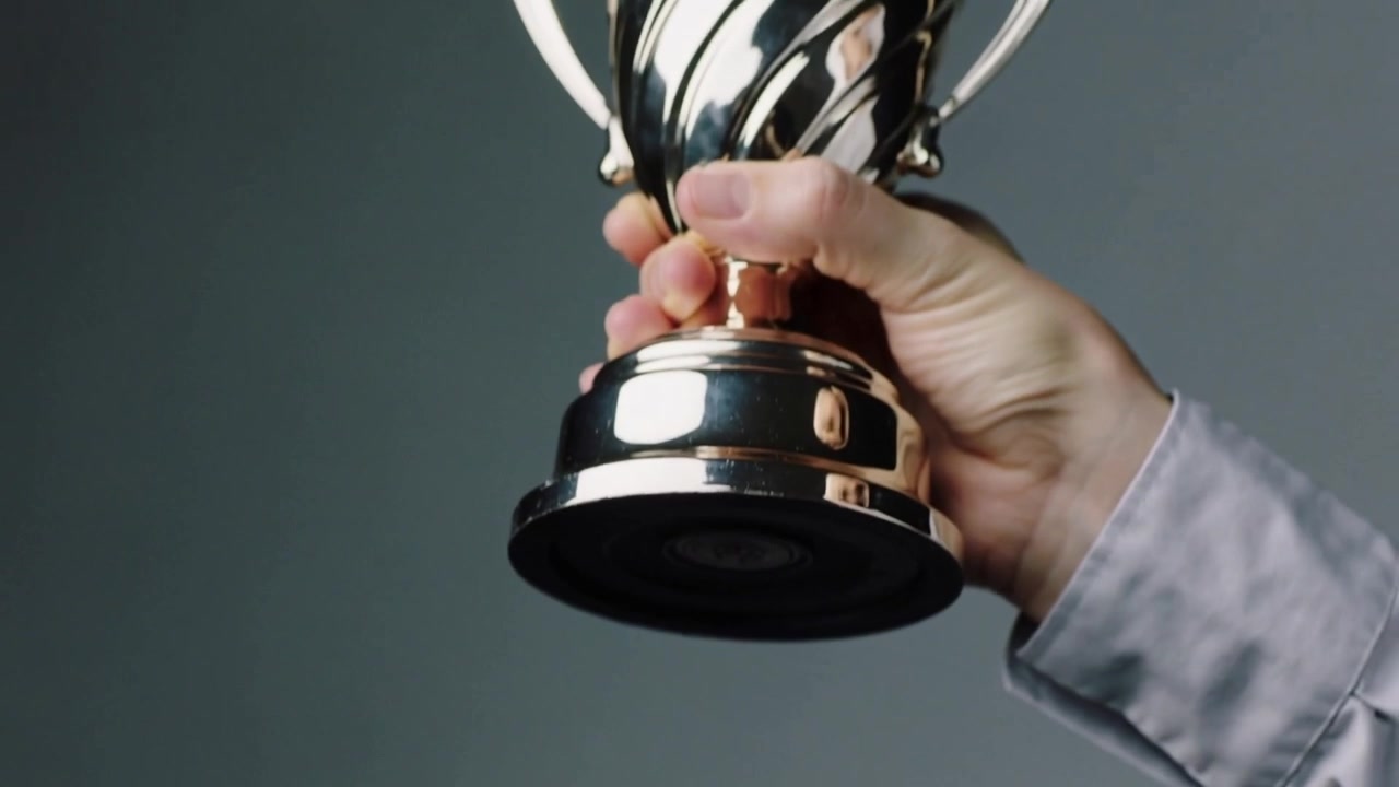 Una mano con una maglia a maniche lunghe chiara che si allunga per sollevare un trofeo d'argento da uno scaffale in una stanza grigia per rimuoverlo.