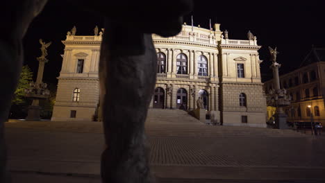 the maginificent concert hall of rudolfinum with bright facade in prague, czechia on an empty square of paved stones at night, during a covid-19 lockdown, shot from behind a statue and its legs