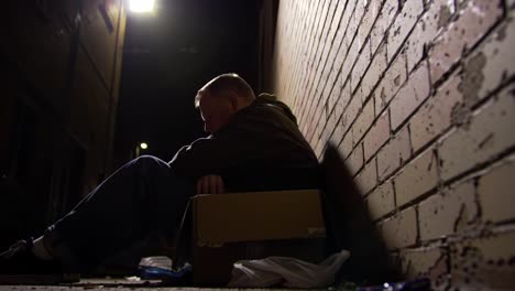 a somber portrayal of a solitary figure seated on a cardboard box in a dimly lit alleyway, illustrating moments of reflection amidst urban challenges and loneliness