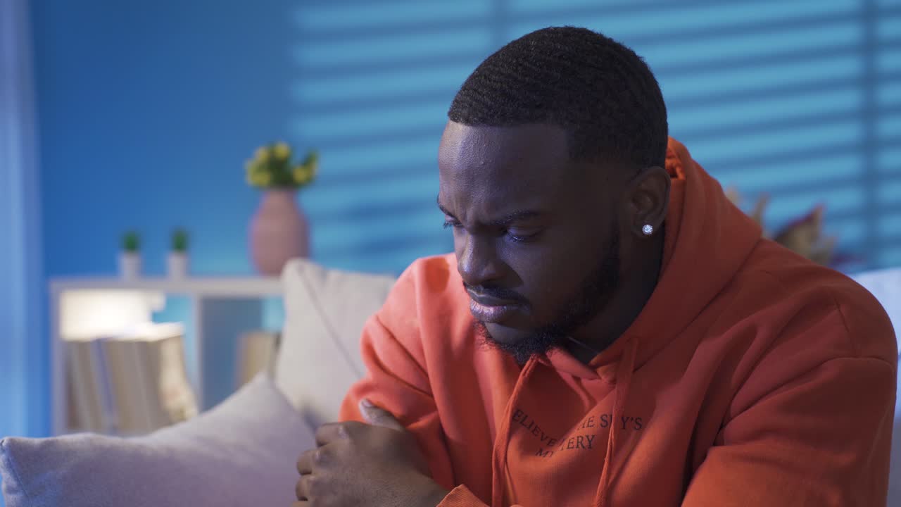 Depressed and anxious black young man sitting alone on sofa at home.