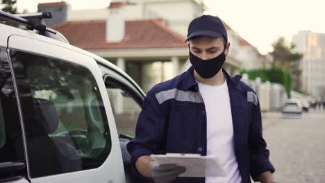 handsome courier going out from the van and takes cardboard box package out of delivery car makes notes in delivery list